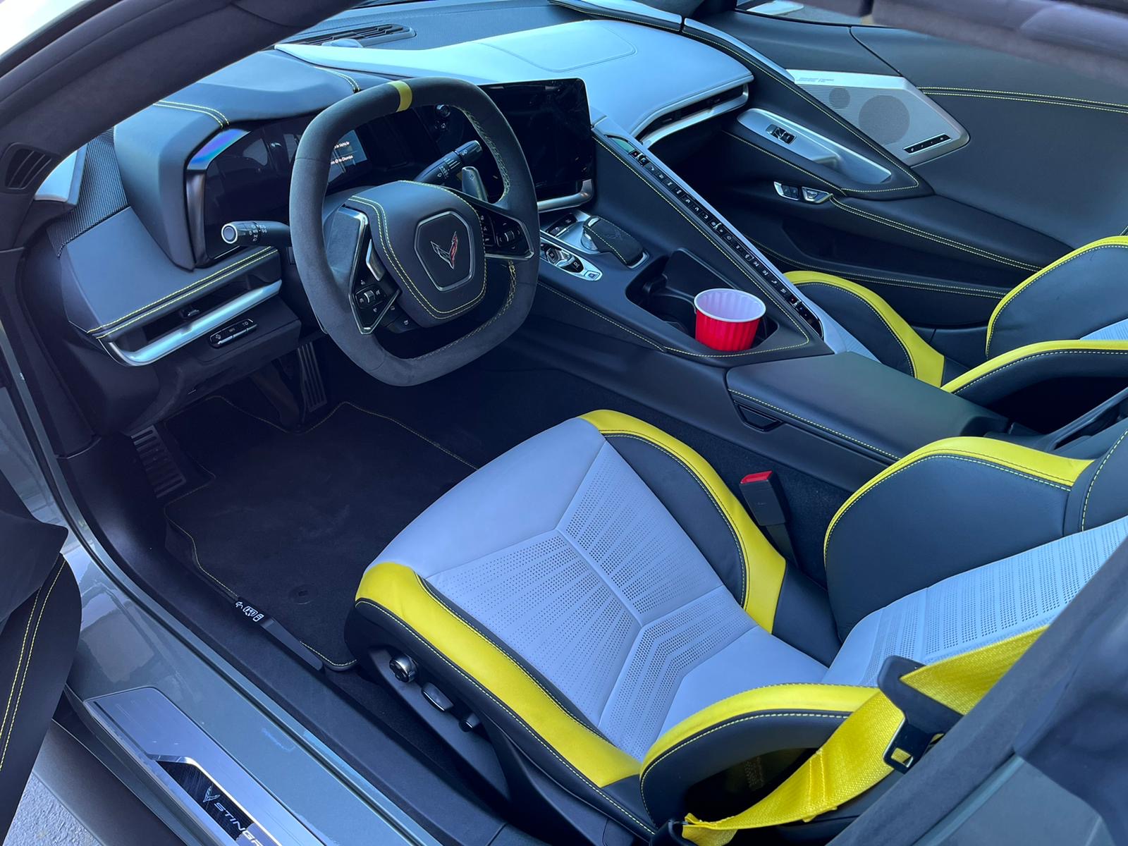 An interior view of a modern sports car cockpit, featuring a two-tone color scheme of black and yellow. The driver's seat has a light gray and yellow perforated leather upholstery with a geometric pattern. The center console has a sleek design with a black and yellow color scheme, featuring a red cup holder. The steering wheel is black with yellow accents and the Corvette logo in the center. The dashboard has a modern, angular design with black leather and yellow trim elements. The interior lighting creates a blue tint throughout the cabin. The gear shifter is positioned in a center console with a yellow accent strip. The passenger seat is partially visible in the frame, matching the driver's seat design. The floor mats are black, and the overall interior has a premium, high-performance aesthetic with attention to detail in the contrast between the black and yellow elements.