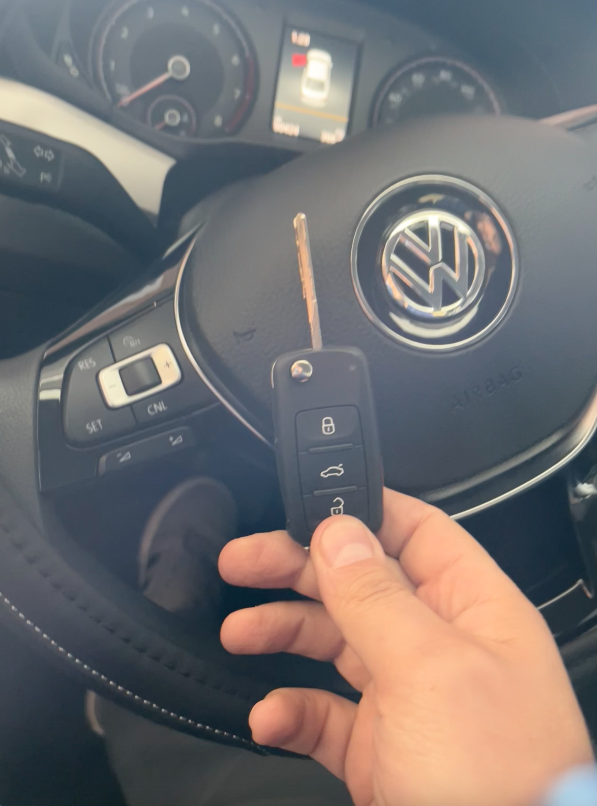A close-up photograph of a hand holding a black Volkswagen car key fob against a dark interior background. The steering wheel of a Volkswagen vehicle is visible in the background, showing the silver VW logo emblem in the center. The steering wheel has black leather upholstery with contrast stitching. On the left side of the steering wheel, there are control buttons with white illuminated symbols. The digital instrument cluster is partially visible in the background, showing red and white illuminated gauges. The image has a warm, slightly desaturated color tone, with the hand's skin tone contrasting against the dark interior elements. The key fob has three buttons visible on its face, with the standard lock, unlock, and trunk release functions. The photograph is taken from inside the driver's side of the vehicle, with the steering wheel slightly angled to the right.