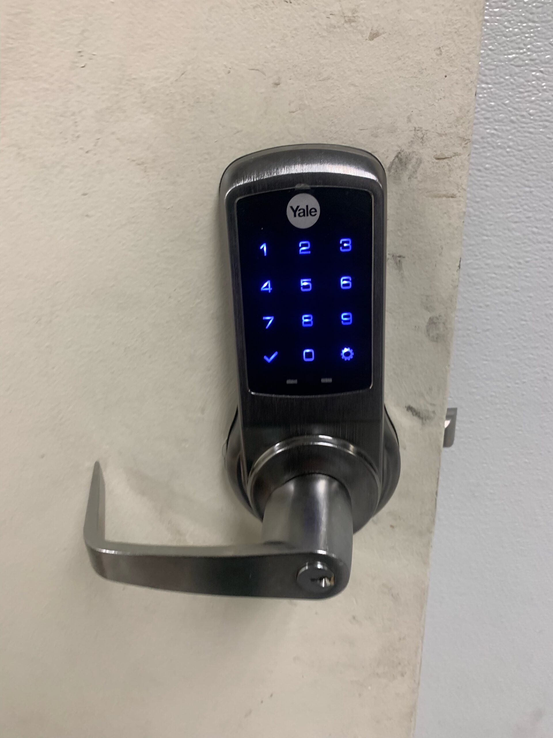 A modern digital door lock system mounted on a light beige wall. The lock has a sleek black and silver design with a digital keypad display. The keypad shows blue illuminated numbers 0-9 arranged in a standard telephone layout. The "Yale" brand logo is in white text at the top of the keypad. The lock features a brushed silver lever handle mounted on a matching silver door preparation. The keypad display is rectangular and housed in a brushed metal casing with rounded edges. The wall behind the lock shows some minor discoloration and wear marks. The image is taken at a slight angle, showing the full length of the lever handle from the lock body to the door surface.
