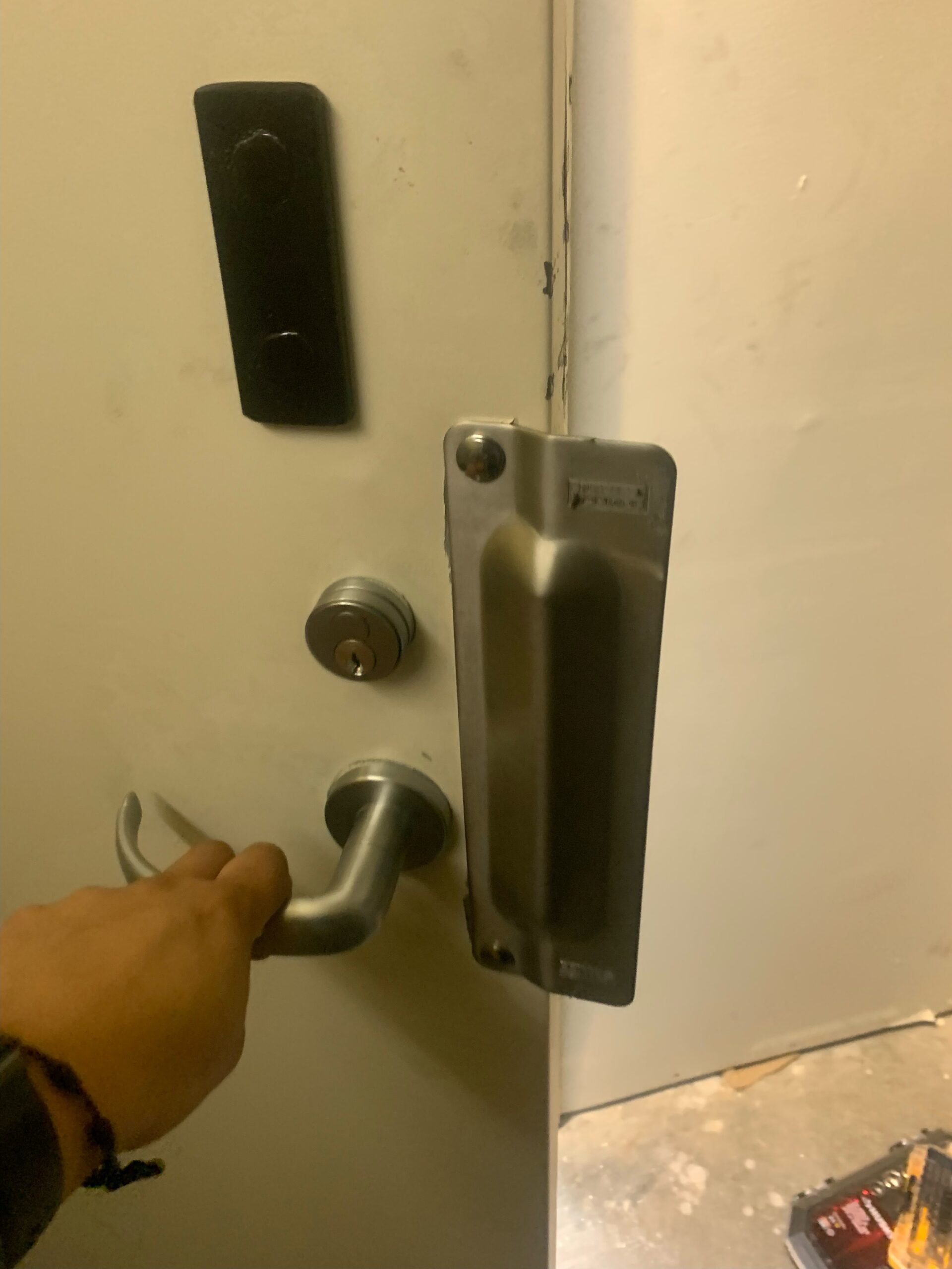 A close-up photograph of a door handle installation process. A hand wearing a black wristband is shown gripping a silver-colored door handle against a white wall. The door handle is mounted on a modern brushed metal door lock mechanism. Above the lock, there is a black rectangular electronic keypad mounted on the wall. The image is taken from a side angle, showing the full length of the door handle from the grip to the mounting plate. The lighting is indoor and warm-toned, casting subtle shadows on the white wall surface. The bottom portion of the image shows a partial view of a concrete floor. The photograph has a slight blur, indicating movement or a quick capture during the installation process.