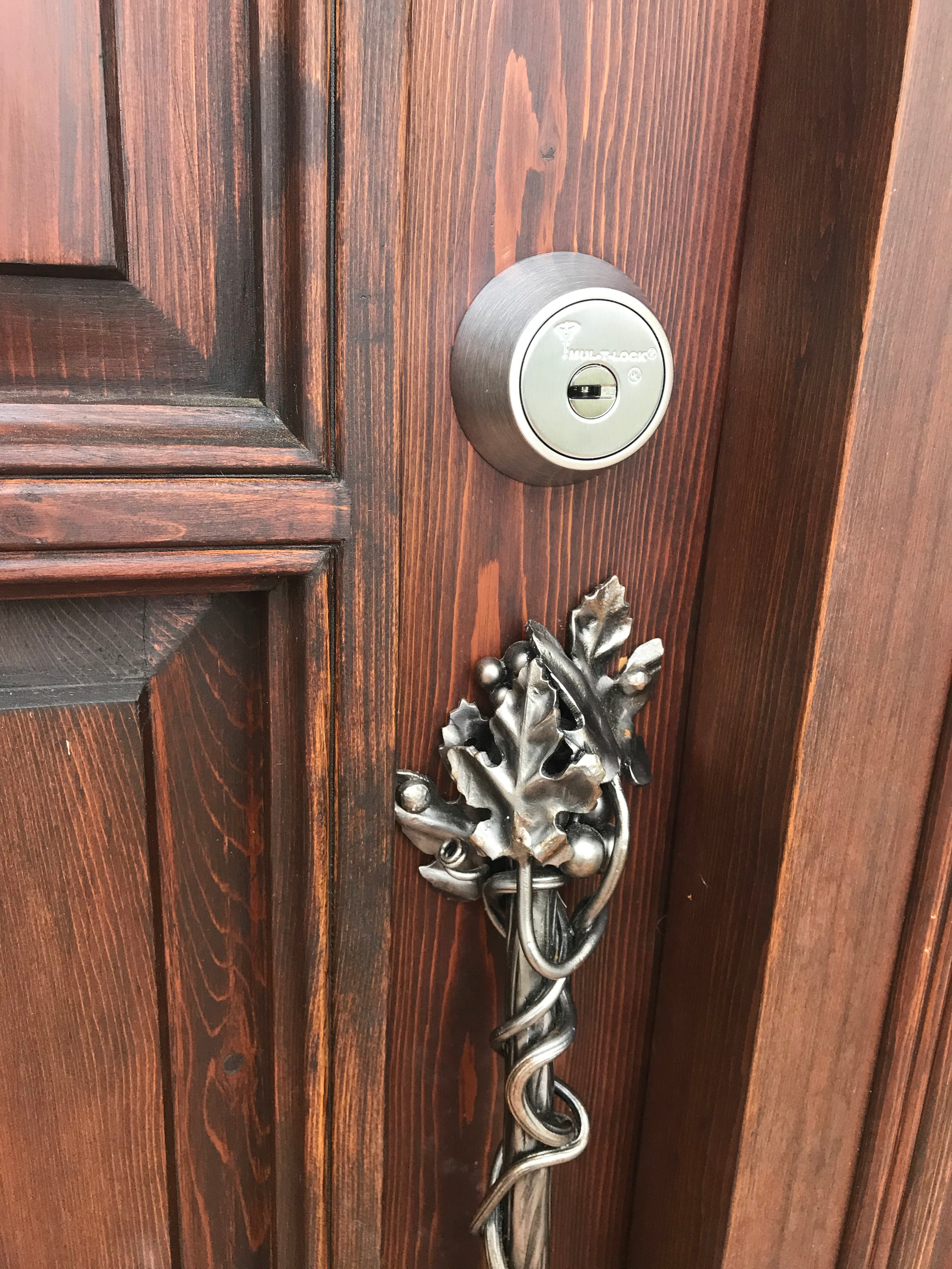 A close-up photograph of an ornate wooden door with rich burgundy-brown stained wood grain texture. The door features a modern brushed nickel circular deadbolt lock and a decorative metal door handle. The handle is crafted in an elaborate vine and leaf design, featuring a large maple leaf at its center with curling vines and smaller leaves extending outward. The metalwork has a dark gunmetal finish that contrasts sharply with the warm wood tones. The door panel has vertical paneling with subtle grooves and raised details. The lighting in the image creates subtle shadows that emphasize the texture of both the wood grain and the metalwork. The photograph is taken at a slight angle, showing the door handle in the lower portion of the frame and the lock mechanism in the upper portion.