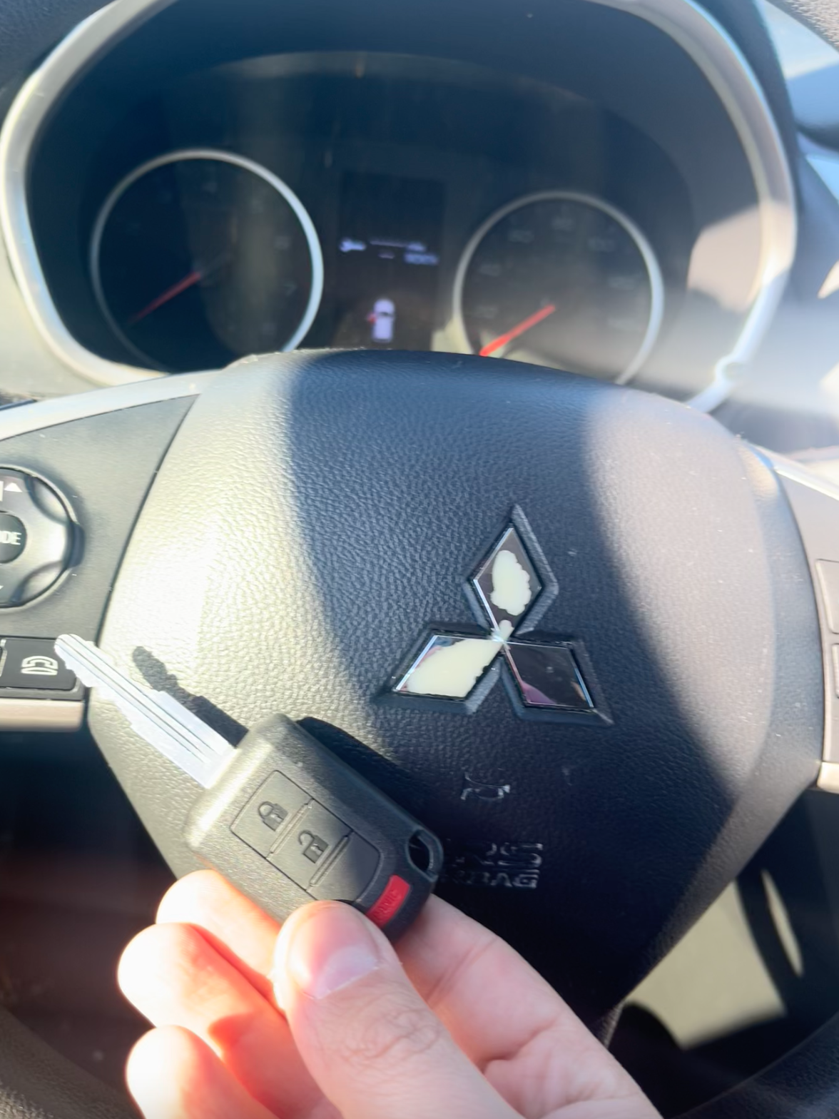A close-up photograph of a Mitsubishi vehicle interior, focusing on the black leather-wrapped steering wheel with the distinctive Mitsubishi diamond logo emblem in silver. A hand is holding a black remote key fob with red accent buttons against the steering wheel. The dashboard is visible in the background, showing two round analog gauges with red needles and a dark interior finish. The steering wheel has silver accent trim around the airbag area and control buttons on the left side. The image is taken from inside the driver's side of the vehicle, with natural lighting coming from outside the vehicle. The leather steering wheel has a textured grip pattern and shows subtle wear marks.