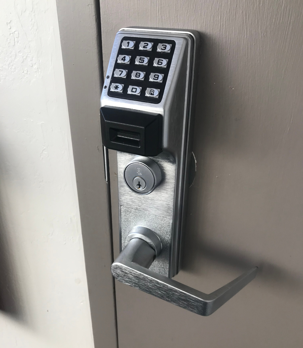 A close-up photograph of a modern electronic keypad door lock mounted on a light gray door. The lock has a brushed metal finish with a sleek digital keypad panel at the top containing white numbers on black buttons. The keypad is protected by a black plastic housing. Below the keypad is a silver circular electronic lock mechanism with a keyhole. The door handle is a modern brushed nickel lever handle mounted on the same plate. The image is shot at a slight angle, showing the door handle on the right side of the frame. The lighting is soft and even, creating subtle shadows around the lock mechanism. The background is a plain light gray wall, slightly out of focus. The photograph has a professional quality with sharp detail on the lock mechanism and clear focus on the keypad and handle.