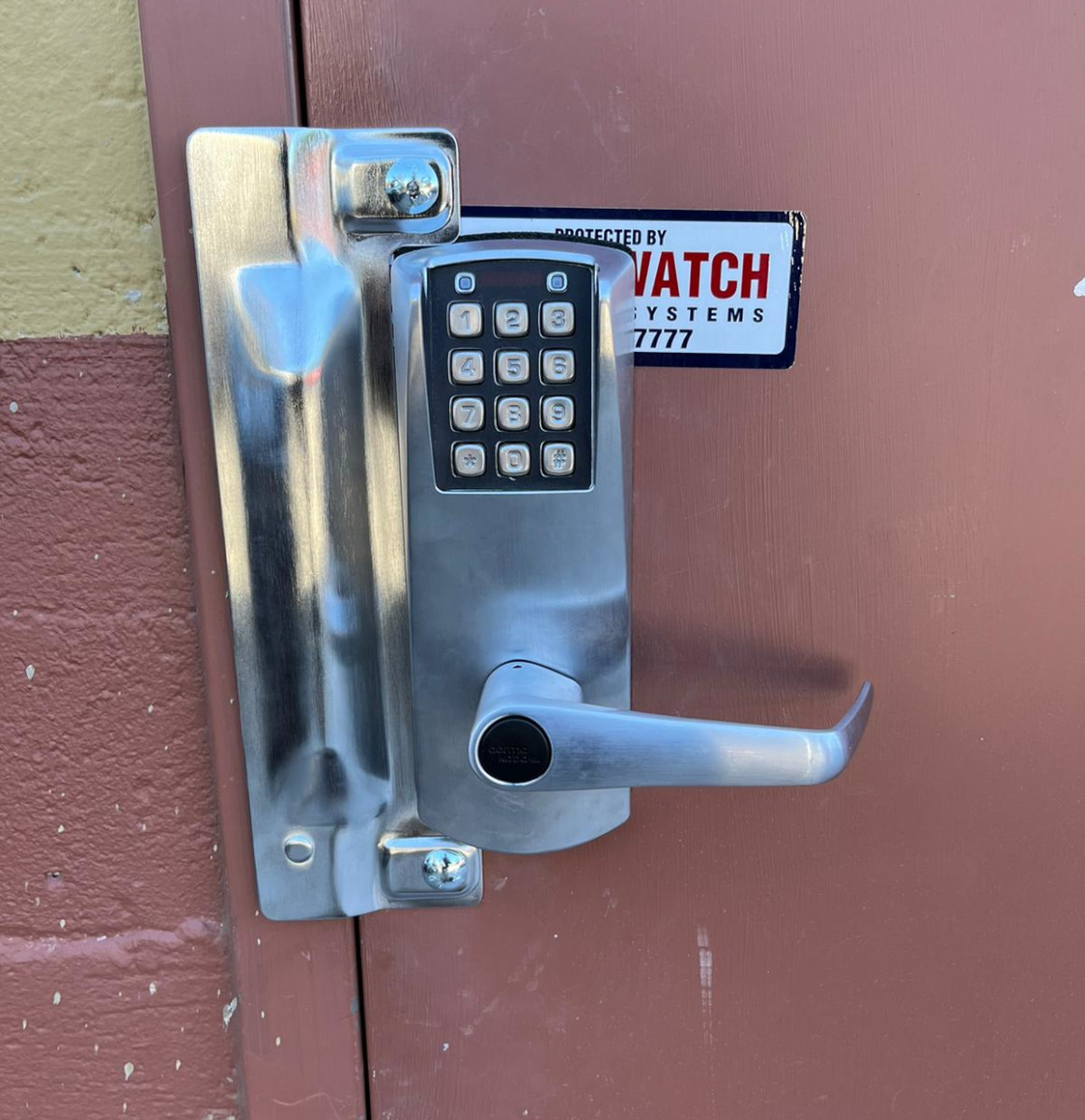 A close-up photograph of a modern digital keypad door lock mounted on a burgundy-colored metal door. The lock has a brushed silver metallic finish with a sleek lever handle design. The keypad panel displays a black numerical keypad with white buttons arranged in a standard 3x4 grid. Above the keypad is a digital display screen. A small white label with black text reading "WATCH SYSTEMS" is mounted next to the lock. The lock mechanism is secured with visible mounting screws and has a protective cover plate. The door surface shows some minor wear and scratches. The image is shot at a slight angle, capturing the metallic sheen of the lock mechanism.