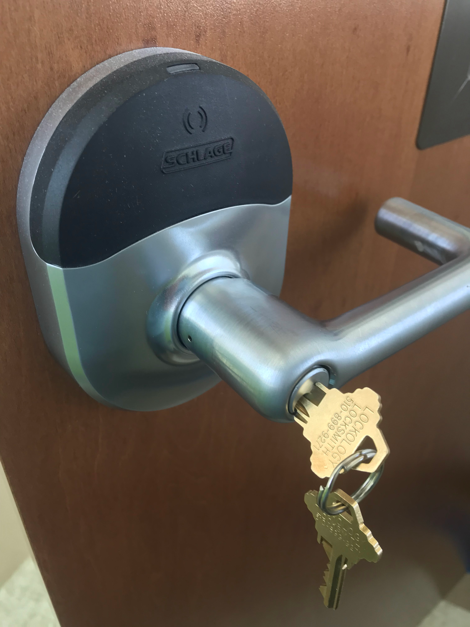 A close-up photograph of a modern Schlage electronic door lock mounted on a dark brown wooden door. The lock has a sleek, curved design with a black upper half and a metallic silver lower half. The Schlage logo is embossed in the black upper portion. A silver-colored door handle is attached to the right side of the lock mechanism. Two brass-colored house keys are inserted into the lock mechanism, hanging down from the keyway. The image is shot from a slight angle, showing the dimensional curves of the lock mechanism against the smooth wooden door surface. The lighting creates subtle reflections on the metallic surfaces of the lock and handle.