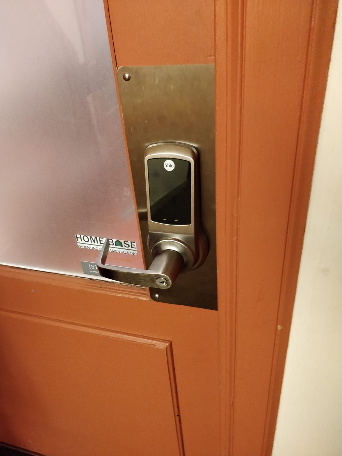 A close-up photograph of a modern electronic door lock mounted on a terracotta-colored wooden door. The lock is a silver digital keypad model with a black display screen and metallic finish. The door handle is a brushed nickel lever. Above the lock is a rectangular brushed metal cover plate in bronze finish. On the left side of the door, there is a small black and white "HOME BASE" security system label. The door frame is painted in the same terracotta color as the door, and white trim is visible along the edges. The image is taken at a slight angle, showing the door handle and lock mechanism in sharp detail against the warm-colored door surface.