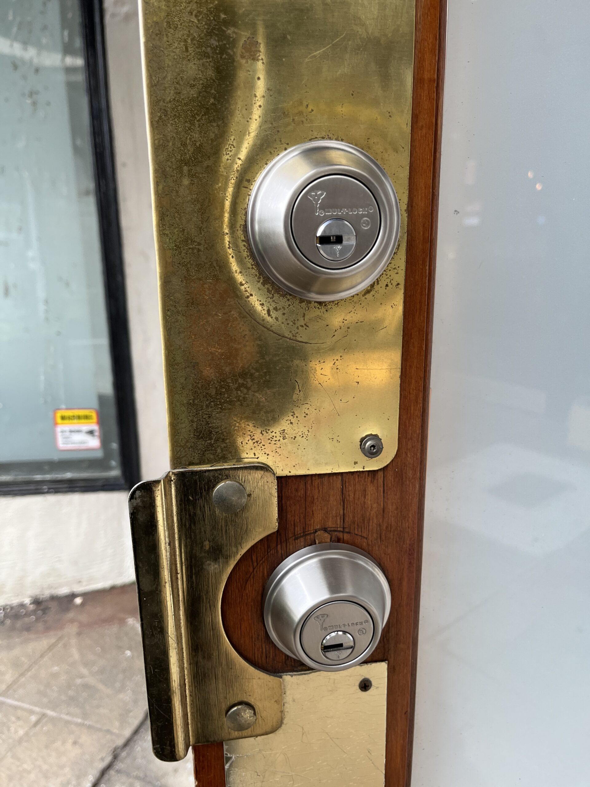 A close-up photograph of a brass door handle and lock mechanism mounted on a wooden door frame. The brass plate shows signs of age and wear with a patina and scratches. Two circular silver-colored deadbolts are mounted on the brass plate - one at the top and one at the bottom. The wooden door frame is visible on the right side of the image, showing a rich brown color and grain pattern. The door handle is partially visible at the bottom of the frame. The image is taken at a slight angle, showing the dimensional depth of the lock mechanism. A small portion of a window frame is visible on the left edge of the image, with a small yellow warning sticker partially visible. The lighting in the image creates subtle reflections on the metallic surfaces, highlighting the worn texture of the brass.