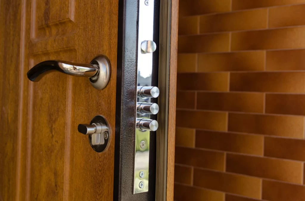 A close-up photograph of a wooden door with a modern security lock system. The door is made of rich brown wood with visible panel details. On the door is a sleek chrome door handle and lock mechanism. The lock system includes a black metal frame with multiple security bolts visible on the right side. The door frame is visible, showing a dark brown trim. In the background, there is a wall with horizontal brown brick tiles. The lighting in the image creates subtle reflections on the chrome hardware and metallic components of the lock system. The image is shot at a slight angle, focusing on the lock mechanism and handle against the warm-toned wood door surface.
