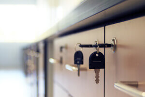 A close-up photograph of two black house keys hanging on a metal key hook against a light beige cabinet surface. The keys are identical and are standard adult-sized house keys with a traditional cross-pattern design. The image has a shallow depth of field, with the background cabinet surface slightly blurred. The lighting is soft and natural, creating subtle shadows behind the keys. The metal key hook is mounted horizontally on the cabinet surface. The cabinet surface has a matte finish and extends beyond the frame on both sides. The photograph has a warm, muted color palette with emphasis on beige and black tones.