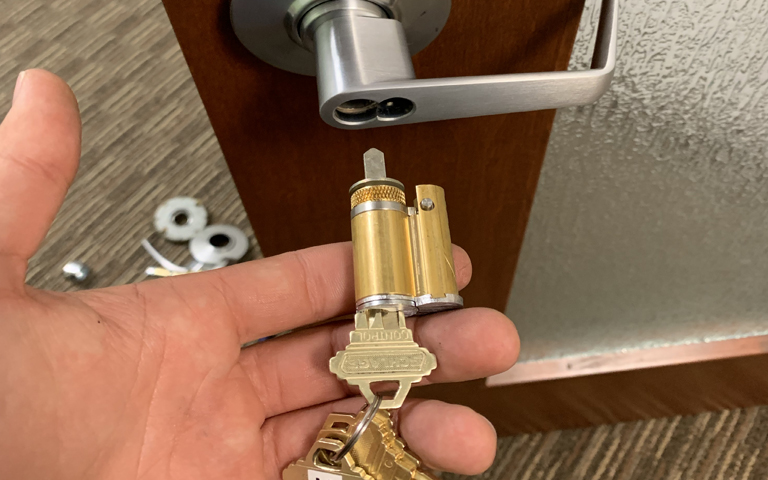 A close-up photograph of a hand holding a brass-colored door lock cylinder and key against a wooden door background. The lock cylinder has a metallic finish with ridged details and a key inserted. The door handle is visible at the top of the frame, showing a modern brushed nickel finish lever handle mounted on a dark brown wooden door. The wall behind the door is textured with a light gray finish. The wooden door has visible wood grain patterns running horizontally across the frame. The hand is positioned from the bottom of the frame, with fingers holding the lock mechanism. The lighting is indoor and even, casting subtle shadows on the wooden surface.