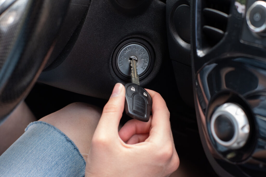 A close-up photograph of a hand inserting a car key into a black plastic ignition switch. The hand is wearing a light blue denim sleeve. The ignition switch is circular and has a metallic silver rim with text engraved on it. The car interior features black dashboard elements and air vents visible in the background. The key is a modern black electronic key fob with metallic accents. The image is shot from a side angle, focusing on the act of starting the vehicle. The lighting is natural and even, highlighting the metallic surfaces of the ignition and key.