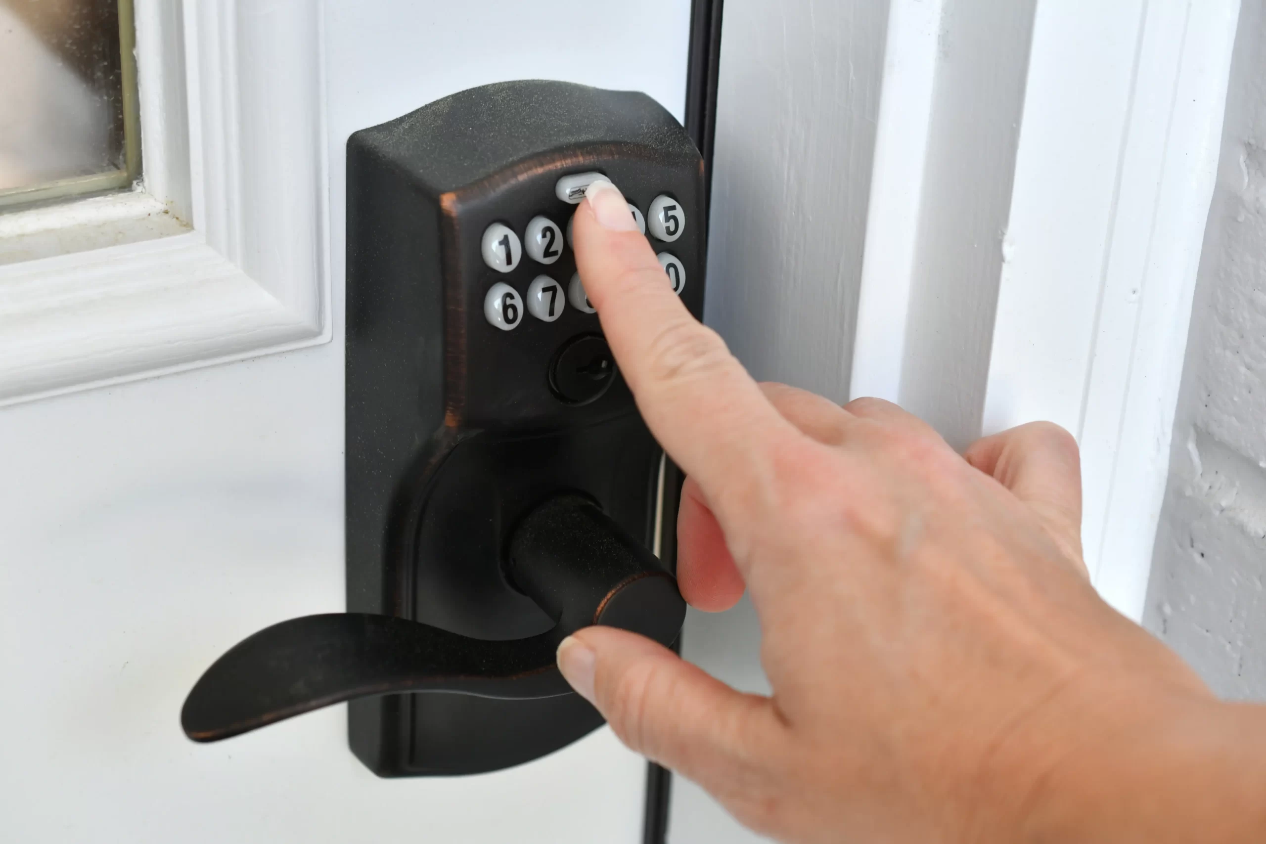 A close-up photograph of a hand entering a code on a black electronic keypad door lock. The lock has a brushed metal finish and features a curved lever handle on the right side. The keypad displays white numbers on a black background, with the numbers 1-0 visible. A single finger is pressing one of the keypad buttons. The lock is mounted on a white door frame, with a small portion of a window visible in the top left corner of the frame. The image is well-lit with natural lighting, creating subtle shadows on the door surface. The hand entering the code is positioned from the right side of the frame, with only the hand and partial forearm visible.