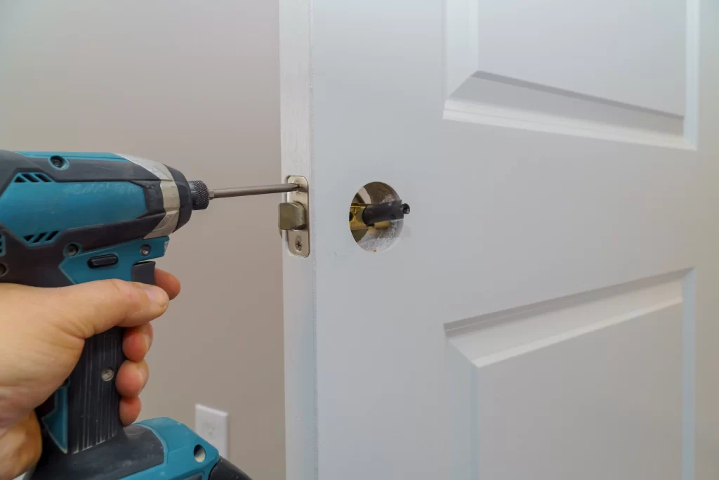 A close-up photograph of a blue and black Makita power drill being used to install a door lock mechanism. The drill is held by a hand entering from the left side of the frame, gripping the turquoise-colored cordless drill with black accents. The drill bit is positioned near a circular hole in a white interior door. The door features a modern three-panel design with raised rectangular details. The door hardware includes a brass-colored lock mechanism installed in a circular hole. The wall behind the door is painted in a light neutral color. The image is well-lit with natural lighting, creating subtle shadows on the door's surface.