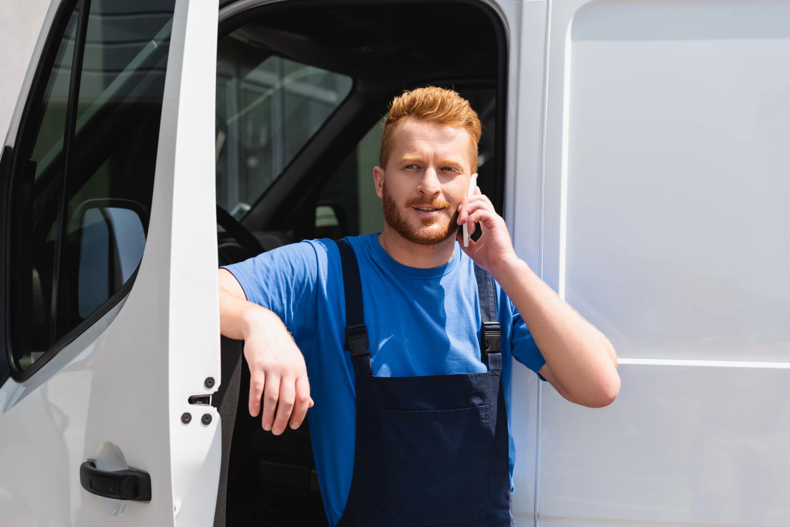 A professional photograph of a caucasian male service worker leaning against a white commercial van with its door open. The worker is wearing a bright blue t-shirt and navy blue overalls, and is engaged in a phone conversation with his right hand. He has vibrant red hair and a well-groomed beard. The van is a service vehicle with a white exterior and visible door handle. The image is shot in natural daylight, creating clear visibility of the subject and vehicle details. The worker's posture is relaxed yet professional, with his left arm resting against the van's frame. The photograph is taken from a slight angle, showing both the side of the vehicle and the worker's upper body. The background is slightly blurred, focusing attention on the subject and vehicle.