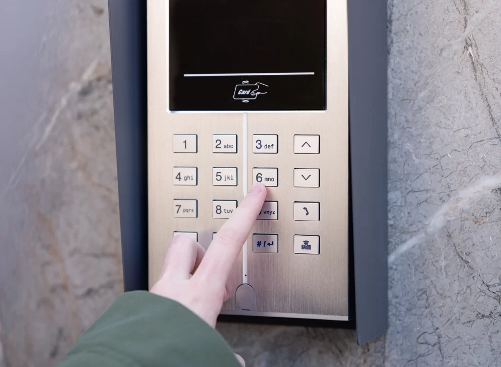 A close-up photograph of a modern intercom keypad mounted on a gray textured wall. The keypad has a metallic silver faceplate with a digital display at the top showing a card payment icon. Below the display is a full numerical keypad with white buttons containing black text showing numbers "0-9", "abc", "def", "ghi", "jkl", "mno", "pqr", "stu", "vwx", "yz", and special characters including "#", "()", and a user icon. A hand wearing a dark green sleeve is pressing the "6" button. The keypad is framed by a dark gray mounting bracket visible at the edges of the frame. The lighting is even and natural, creating subtle reflections on the metallic surface of the keypad.