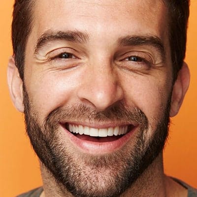 A close-up portrait photograph of a middle-aged man against an orange background. He has a warm, genuine smile showing white teeth, and well-groomed dark brown facial hair including a neatly trimmed beard and mustache. His dark brown hair is styled short and slightly swept to the side. The lighting is professional studio-style, creating soft shadows and highlighting the natural contours of his face. His eyes are crinkled in a genuine expression of joy. The image is cropped to focus on the face from just below the ears to the top of the head. The skin texture is clear and detailed, showing natural aging characteristics. The photograph has a warm color temperature that complements the orange backdrop.