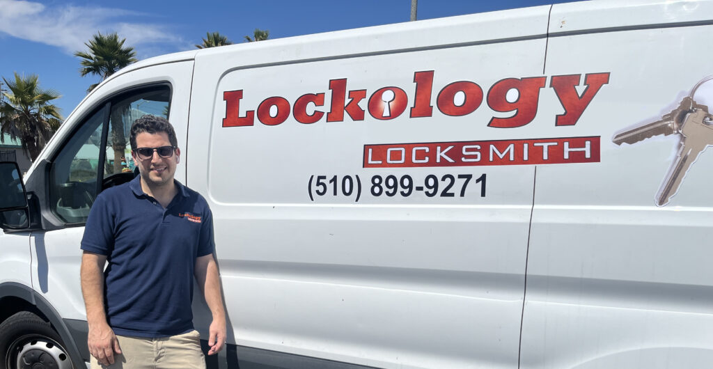 A professional business photo of a male locksmith standing next to a white commercial van. The van has red text reading "Lockology" and "LOCKSMITH" on its side, along with a phone number "(510) 899-9271". The van is a cargo van with a white exterior and black trim. The background shows a bright blue sky with white clouds, and palm trees are visible in the distance. The person is wearing a navy blue polo shirt, khaki pants, and sunglasses. The van's side panel features a small key icon in red. The photo is taken during daytime with natural sunlight illuminating the scene. The composition is centered with the person positioned on the left side of the frame, with the van taking up most of the right side of the image.