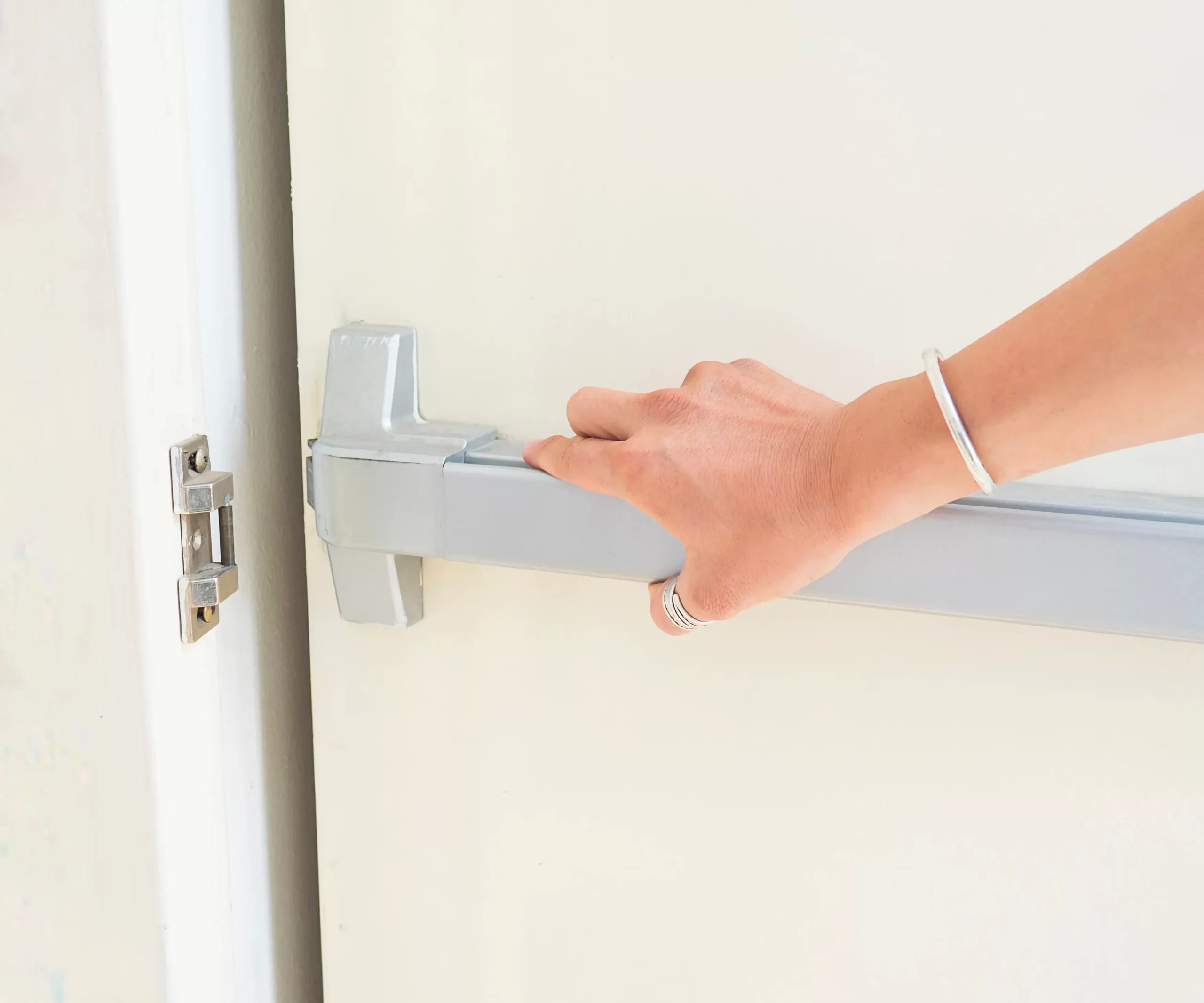 A close-up photograph of a hand opening a white door with a modern gray metal door handle. The hand is wearing a white bracelet and a silver ring. The door handle is mounted on a white door frame with visible hinges. The image is cropped to focus on the hand and handle, with the rest of the door and wall visible but out of frame. The lighting is bright and even, creating a clean, minimalist aesthetic. The door handle is a push bar style, mounted horizontally across the door face. The surface of the door is smooth and painted in a bright white color.