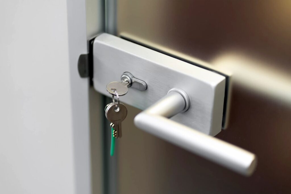 A close-up photograph of a modern brushed aluminum door handle and lock mechanism. The door handle is mounted on a glass panel with a metal frame. The handle is sleek and minimalist in design, with a long horizontal bar in brushed silver finish. A key with a green keychain tag is inserted into the lock mechanism, which is mounted on a rectangular brushed aluminum plate. The lock mechanism has a cylindrical keyhole and a small release button. The background is softly blurred, creating a shallow depth of field effect. The lighting is soft and even, highlighting the metallic surfaces and creating subtle reflections on the glass and metal surfaces. The image is cropped to focus on the door handle and lock mechanism, with the handle positioned slightly off-center in the frame.
